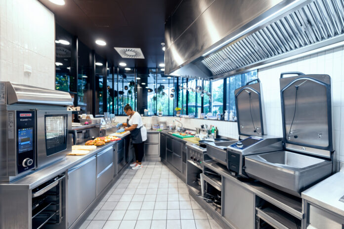 RATIONAL_Reference_Cafe_Mokka_08219_1024992 Commercial kitchen with stainless steel appliances; a chef prepares sandwiches on the counter.