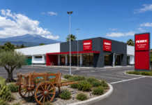 Exterior view of a Penny supermarket in San Giovanni La Punta with red signs and a dark gray facade; a colorful decorative wagon sits in the landscaped island in the foreground; mountains in the distance.