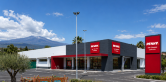 PENNY rafforza la presenza nel Catanese con un nuovo punto vendita Exterior view of a Penny supermarket in San Giovanni La Punta with red signs and a dark gray facade; a colorful decorative wagon sits in the landscaped island in the foreground; mountains in the distance.