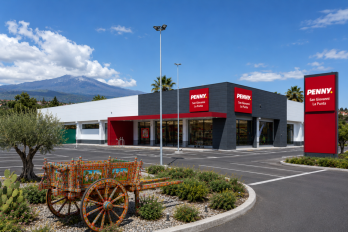 SGLP Exterior view of a Penny supermarket in San Giovanni La Punta with red signs and a dark gray facade; a colorful decorative wagon sits in the landscaped island in the foreground; mountains in the distance.