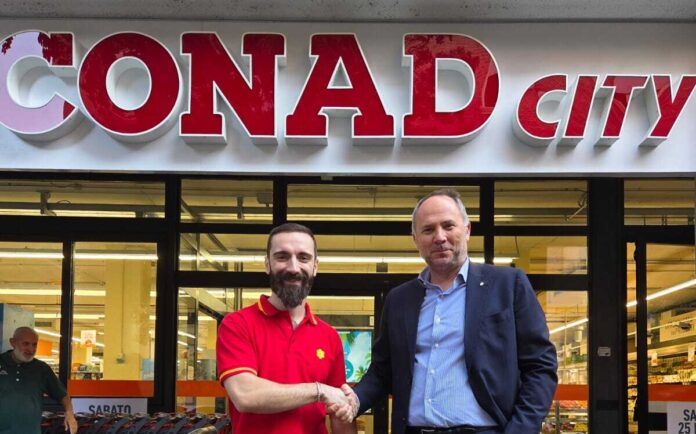 Two men shake hands in front of a CONAD City supermarket storefront