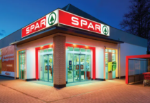 Exterior view of a SPAR supermarket at dusk with glass automatic doors, red signage, and a brick pillar.