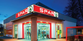 Spar International raggiunge 50,9 miliardi di fatturato e rafforza la rete globale Exterior view of a SPAR supermarket at dusk with glass automatic doors, red signage, and a brick pillar.