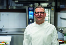 RATIONAL valorizza la cucina di pesce tra tradizione, tecnologia e sostenibilità Male chef wearing a white coat and glasses standing in a stainless steel kitchen, smiling at the camera.