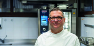 Male chef wearing a white coat and glasses standing in a stainless steel kitchen, smiling at the camera.