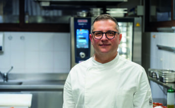 Male chef wearing a white coat and glasses standing in a stainless steel kitchen, smiling at the camera.