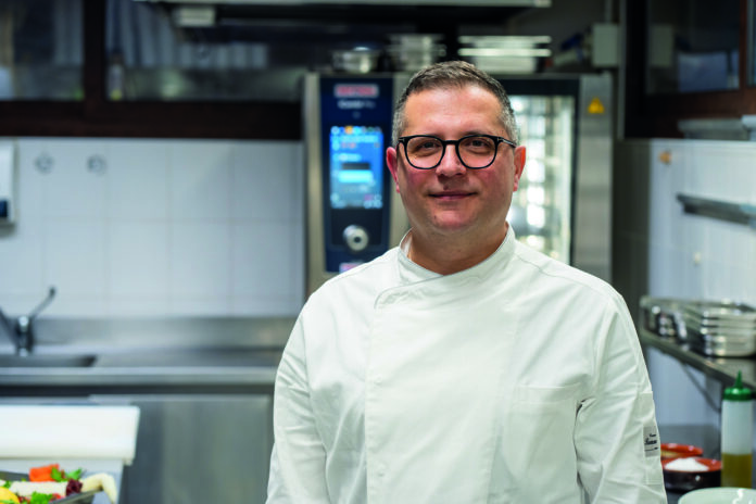 Taverna del Borgo_2 Male chef wearing a white coat and glasses standing in a stainless steel kitchen, smiling at the camera.