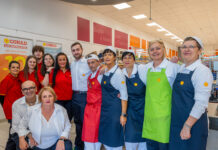 Group of CONAD supermarket staff posing for a photo, wearing red shirts and blue or green aprons with yellow flowers.