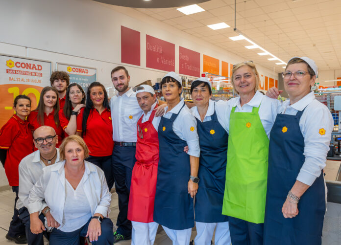 Team Conad San Martino in Rio Group of CONAD supermarket staff posing for a photo, wearing red shirts and blue or green aprons with yellow flowers.