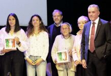 Group of six adults on a stage posing for a photo, two women in front hold framed pictures.