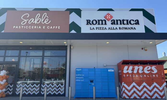 Unes Vimodrone (1) Front of a shopping plaza with Sablé Pasticceria e Caffè and RomAntica La Pizza alla Romana signs, glass doors, and blue Amazon lockers beside red parcel lockers; balloons on the left.