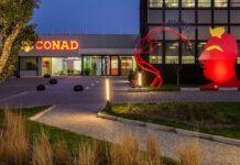 Conad City, nuovo punto vendita nel Fiorentino con focus su freschi e localismo Night exterior of a modern building with a lit CONAD sign and a large red abstract sculpture in the lawn, at dusk.