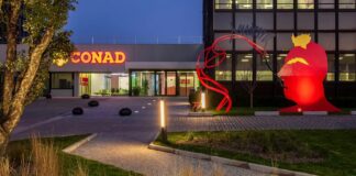 Night exterior of a modern building with a lit CONAD sign and a large red abstract sculpture in the lawn, at dusk.