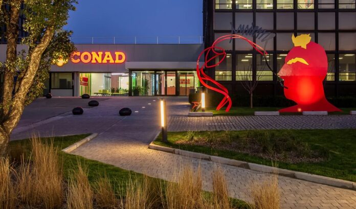 all Night exterior of a modern building with a lit CONAD sign and a large red abstract sculpture in the lawn, at dusk.