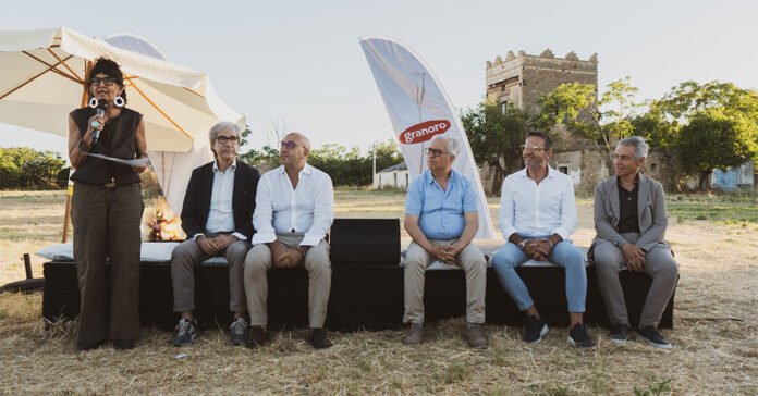 Outdoor panel discussion with six people seated on a black stage; a woman on the left speaks into a microphone, Granoro banner in the background.