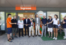 Etruria Retail rafforza la presenza in Toscana con il nuovo Famila Market Group of people at a ribbon-cutting ceremony outside a store entrance labeled 'Ingresso' with colorful balloons and a speaker in a suit holding scissors/ microphone.