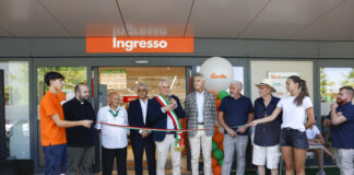 Group of people at a ribbon-cutting ceremony outside a store entrance labeled 'Ingresso' with colorful balloons and a speaker in a suit holding scissors/ microphone.