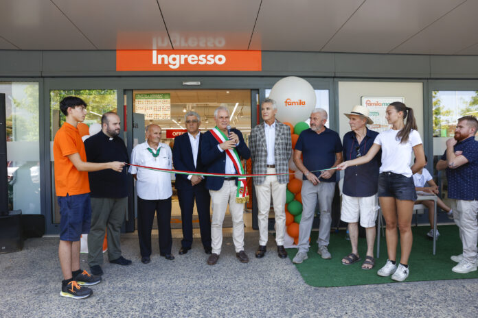 Group of people at a ribbon-cutting ceremony outside a store entrance labeled 'Ingresso' with colorful balloons and a speaker in a suit holding scissors/ microphone.