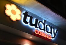 Tuday Conad arriva a Milano nel punto vendita di via Buonarroti Nighttime exterior sign for a Conad supermarket with a yellow-orange flower logo and the word 'CONAD' in red letters.