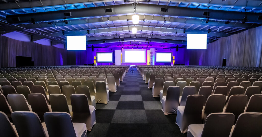Rows of empty chairs in the large conference hall for corporate convention or lecture Large conference hall with rows of white-covered chairs facing a lit stage and multiple screens on the front wall.