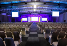 Eventi corporate, perché anche la location costruisce posizionamento Large conference hall with rows of white-covered chairs facing a lit stage and multiple screens on the front wall.