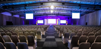 Large conference hall with rows of white-covered chairs facing a lit stage and multiple screens on the front wall.