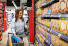 Woman shopping in a supermarket aisle, pushing a cart and looking at her phone while holding a cereal box.