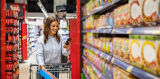Retail media nella GDO: nuova leva di marginalità o rischio di saturare il cliente? Woman shopping in a supermarket aisle, pushing a cart and looking at her phone while holding a cereal box.