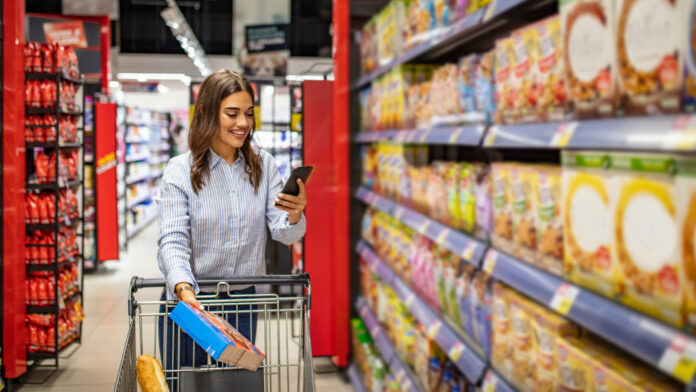 Woman shopping in a supermarket aisle, pushing a cart and looking at her phone while holding a cereal box.
