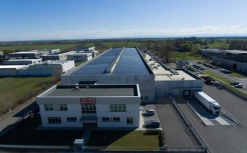 Comal Salumi rafforza il dialogo con retail e filiera attraverso il primo Bilancio ESG Aerial view of a large industrial facility with solar panels on the roof and delivery trucks at a loading dock.