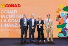 Conad chiude il 2025 a 21,7 miliardi e presenta un piano investimenti da 2,3 miliardi Four professionals stand on a stage in front of a large orange CONAD backdrop with Italian text and logo, posing for a photo.
