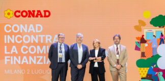 Conad chiude il 2025 a 21,7 miliardi e presenta un piano investimenti da 2,3 miliardi Four professionals stand on a stage in front of a large orange CONAD backdrop with Italian text and logo, posing for a photo.