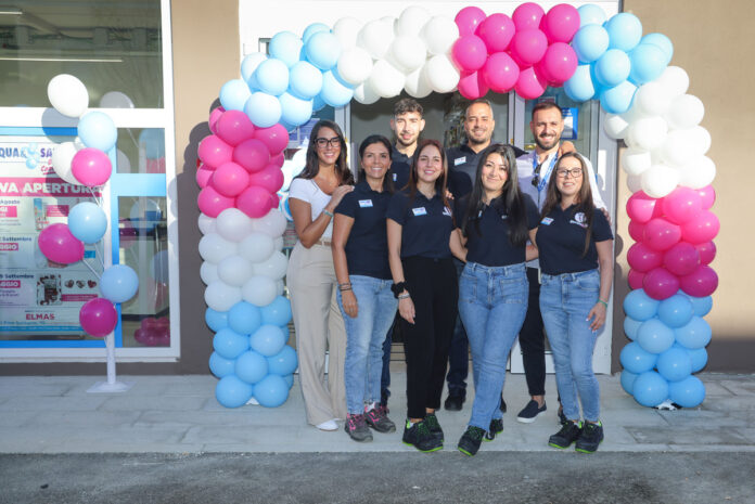 Nine staff members in navy shirts pose under a colorful balloon arch at the store entrance, smiling for a group photo.