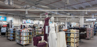Store interior with mannequins wearing maroon and white outfits in the center, surrounded by shelves of folded clothes and a 'Womens' sign in the background.