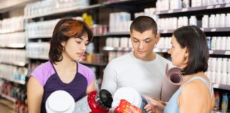 High protein, lo scaffale entra nella fase della selezione Three customers in a supplement store compare large protein powder containers. They examine labels and discuss their choices in front of stocked shelves.