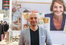 Man in a gray blazer at a trade show booth with product jars and boxes, backdrop of a smiling woman photo behind him.
