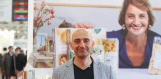 Fumaiolo: «Il freezer può diventare il nuovo spazio premium della pasta fresca» Man in a gray blazer at a trade show booth with product jars and boxes, backdrop of a smiling woman photo behind him.