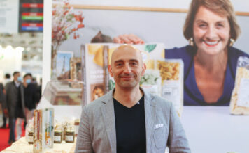 Fumaiolo: «Il freezer può diventare il nuovo spazio premium della pasta fresca» Man in a gray blazer at a trade show booth with product jars and boxes, backdrop of a smiling woman photo behind him.