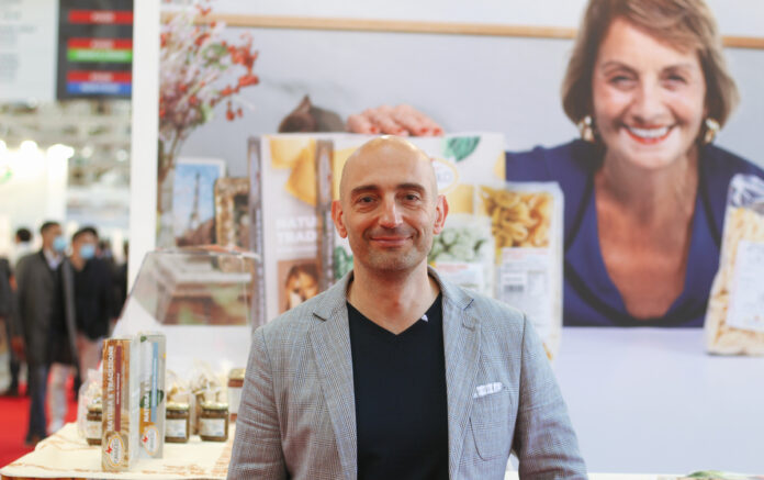 3B0A82AF-159B-43AC-BA9B-3B10FD01 Man in a gray blazer at a trade show booth with product jars and boxes, backdrop of a smiling woman photo behind him.
