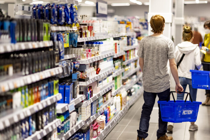 ACTION_repertorio_6 Shoppers with blue baskets walk down an aisle stocked with cosmetics and personal care products in a bright store.