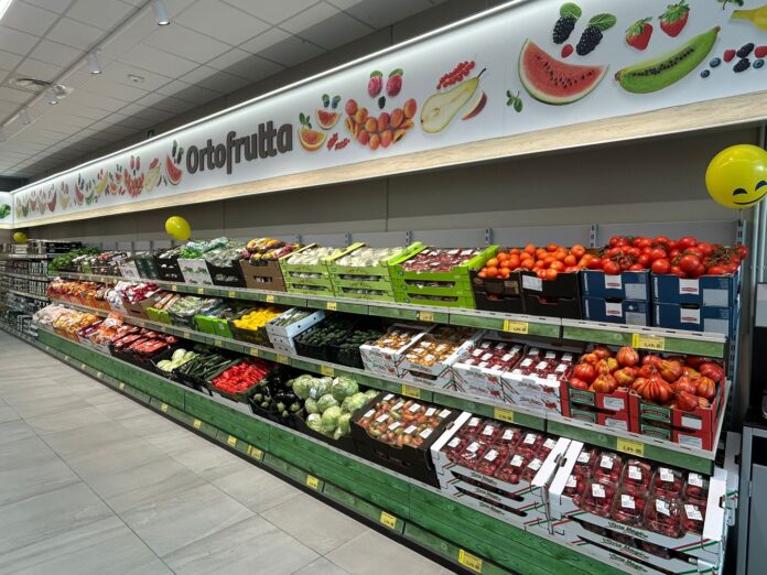 Apertura Villa Cortese Supermarket produce aisle with a colorful fruit banner reading 'Ortofrutta' and crates of tomatoes and other fruit below.