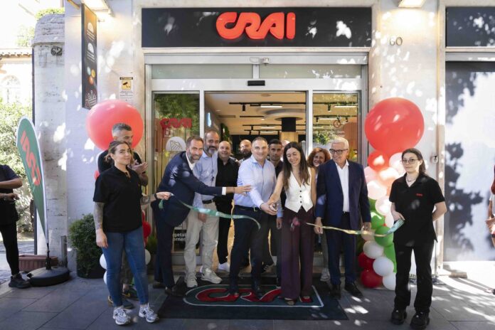 Crai_02 Group of people cutting a ribbon at a CRAI storefront opening, surrounded by balloons and onlookers outside the shop.