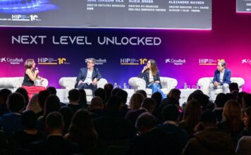 Panel discussion on stage with four speakers seated in white armchairs under a large 'NEXT LEVEL UNLOCKED' banner; audience visible in foreground.