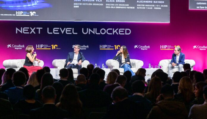 HIP Italia riunira? i protagonisti del settore della pizza Panel discussion on stage with four speakers seated in white armchairs under a large 'NEXT LEVEL UNLOCKED' banner; audience visible in foreground.
