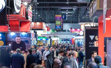 Crowded trade show hall with colorful exhibitor booths, hanging signs, and many attendees walking between aisles under bright ceiling lights.