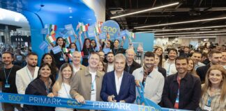 Primark apre a Pompei e raggiunge quota 500 store a livello globale Group of diverse people at a ribbon-cutting for Primark's 500th store, holding a blue 'PRIMARK' ribbon and small flags.