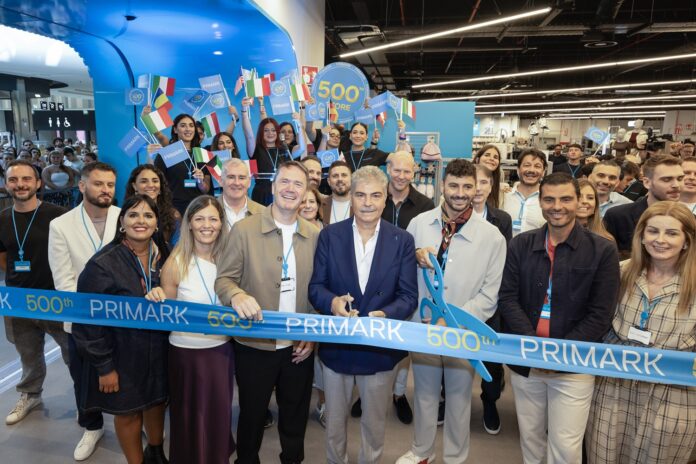 La Cartiera Primark Store 500 NSO Group of diverse people at a ribbon-cutting for Primark's 500th store, holding a blue 'PRIMARK' ribbon and small flags.
