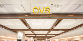 OVS apre a Dubai il più grande flagship store del Gruppo Bright clothing store interior with mannequins displaying outfits, circular display platforms, and racks of apparel under a white ceiling with wooden beams at the top of the aisle.