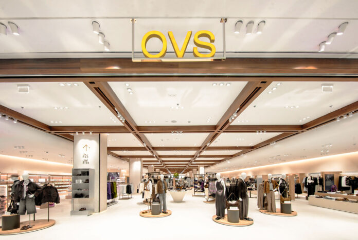 Bright clothing store interior with mannequins displaying outfits, circular display platforms, and racks of apparel under a white ceiling with wooden beams at the top of the aisle.