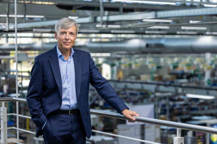 Executive in a navy suit standing on a mezzanine railing overlooking a manufacturing floor.
